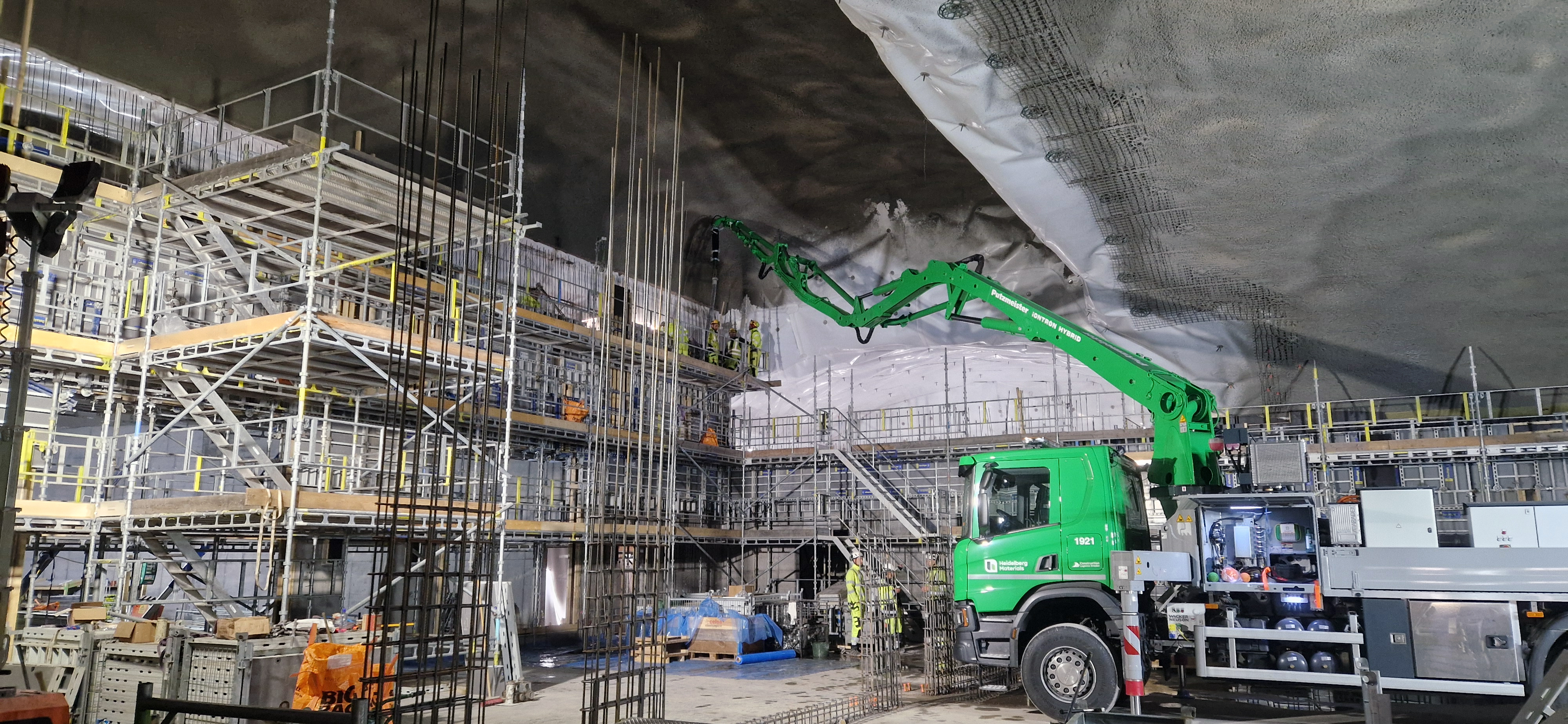 Byggarbetsplats inuti en tunnel med höga byggställningar till vänster, en grön betongbil med lång arm som pumpar betong på tunnelns tak till höger, och byggarbetare runtom som utför arbete.
