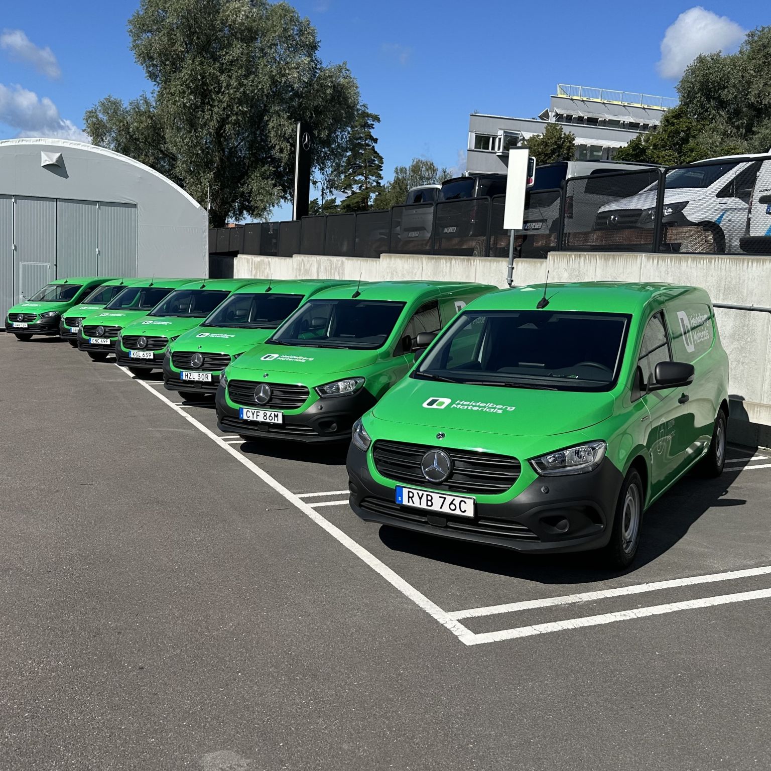 Gröna leveransskåpbilar parkerade i rad på en asfalterad parkeringsplats bredvid en vit byggnad, med träd och en klarblå himmel i bakgrunden.