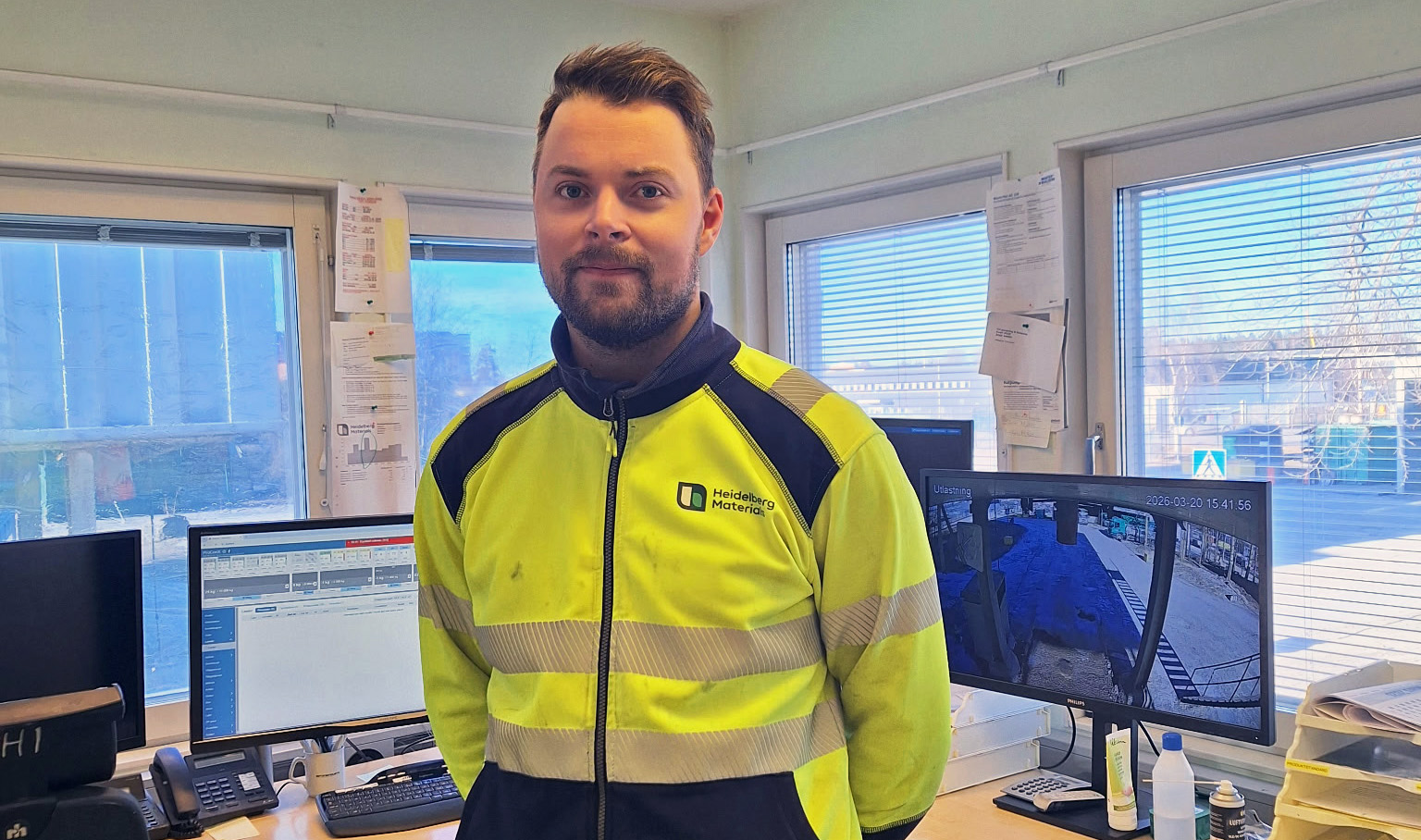 Person i arbetskläder står i ett kontor med flera datorskärmar och fönster i bakgrunden.