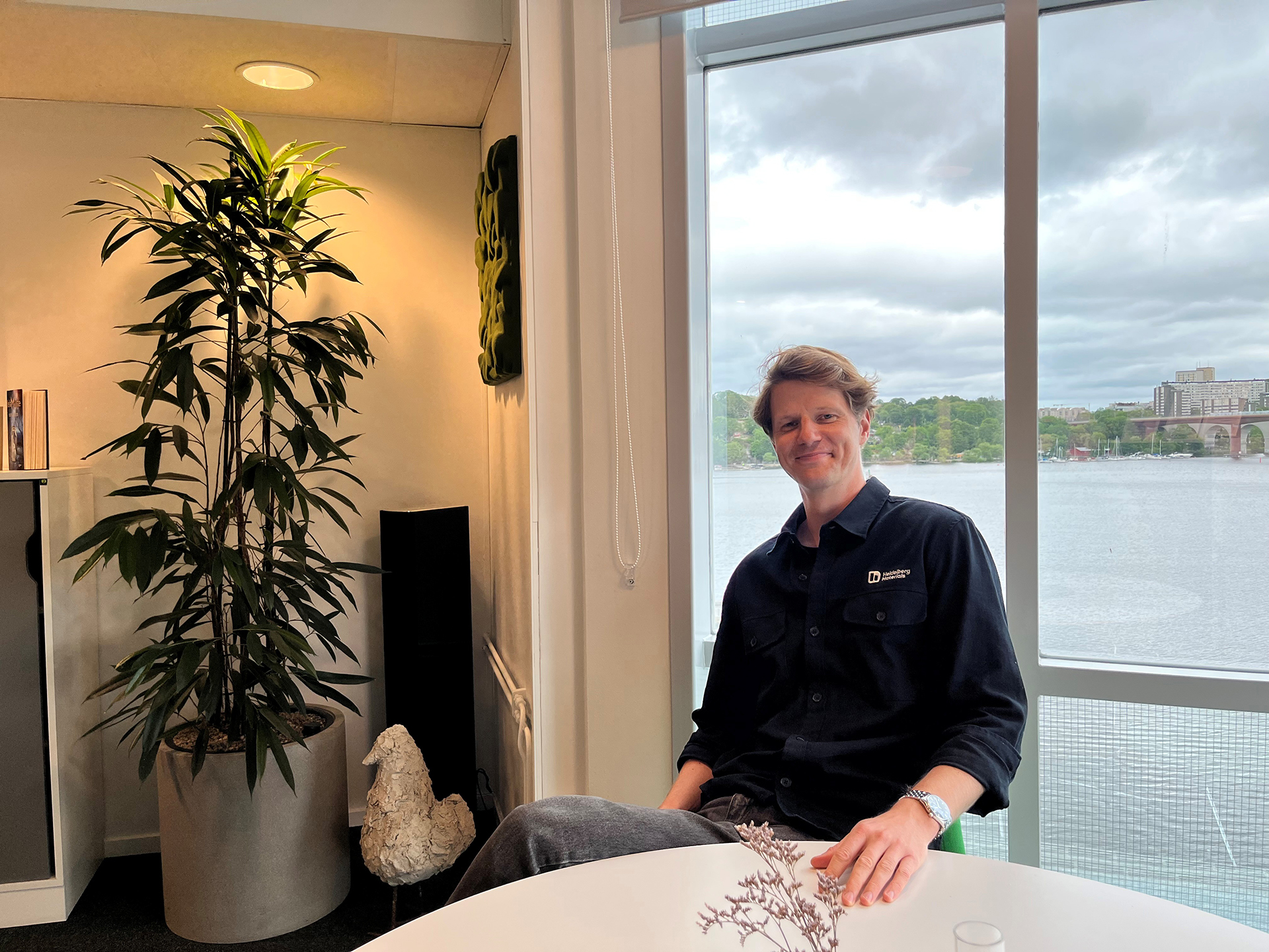 En person sitter vid ett bord i ett rum med stora fönster och utsikt över vatten.