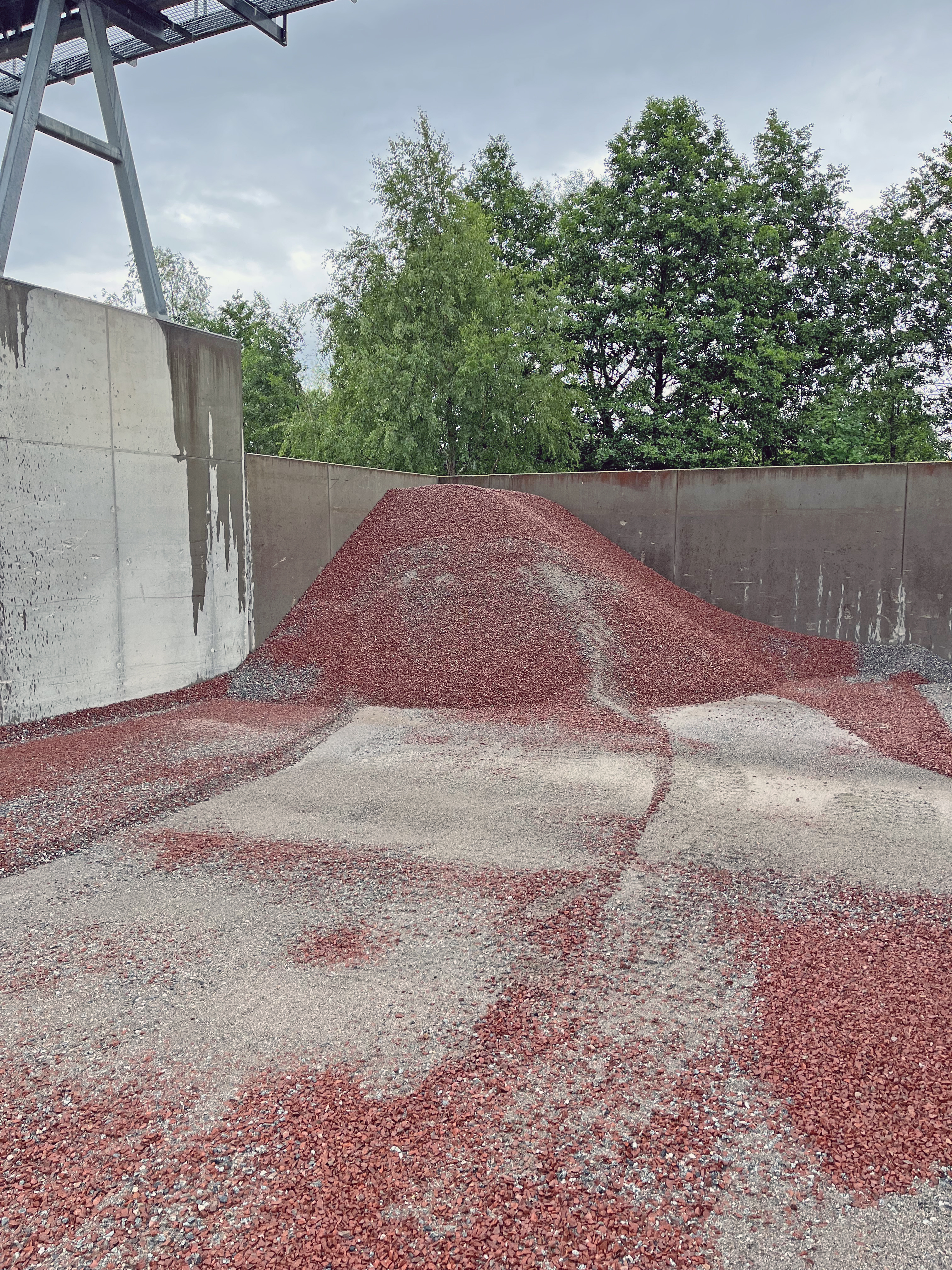 En hög med röda grusmaterial ligger i en betongbehållare med gröna träd i bakgrunden.