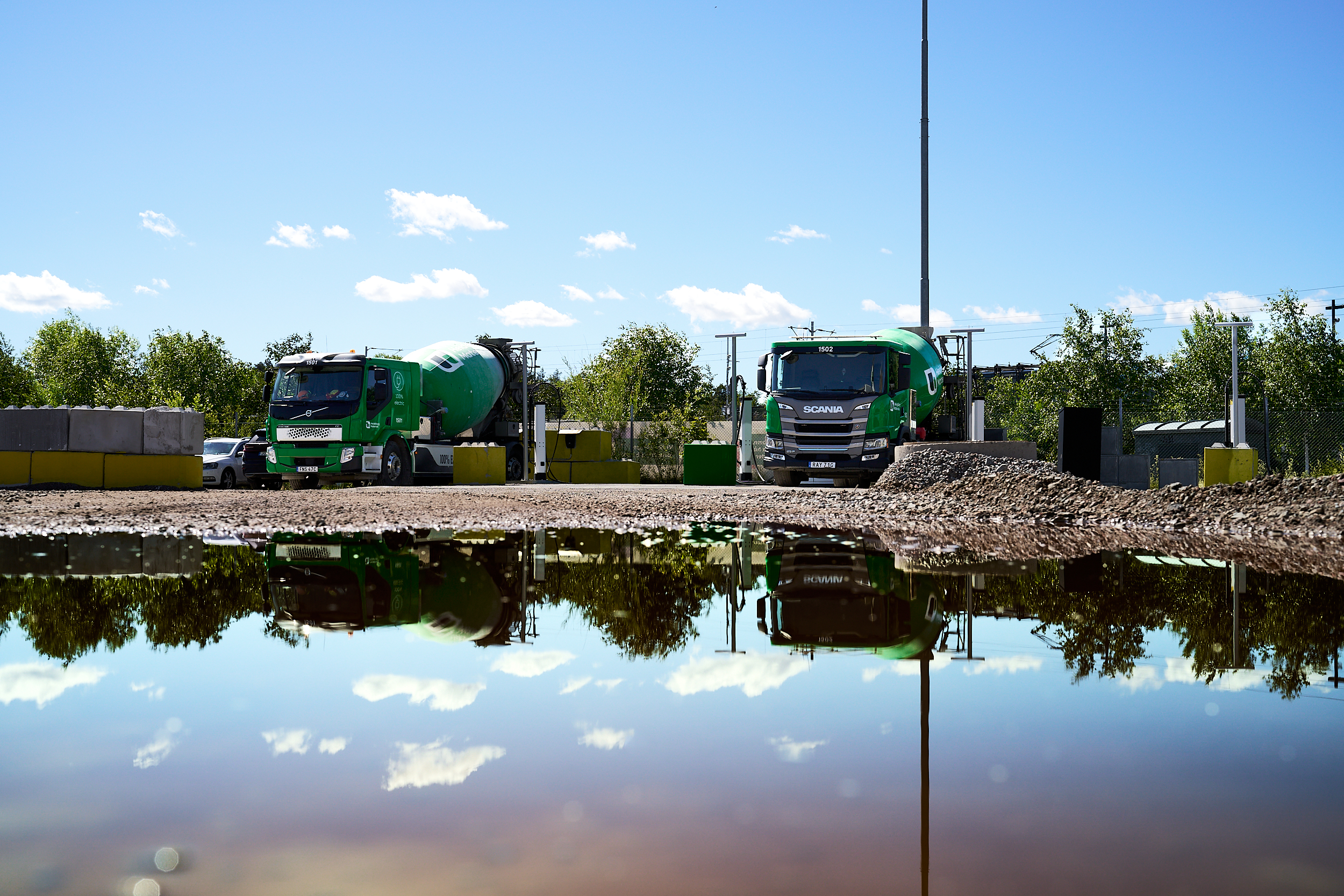 Gröna betongbilar står vid en industriell anläggning med vattenpölar i förgrunden.