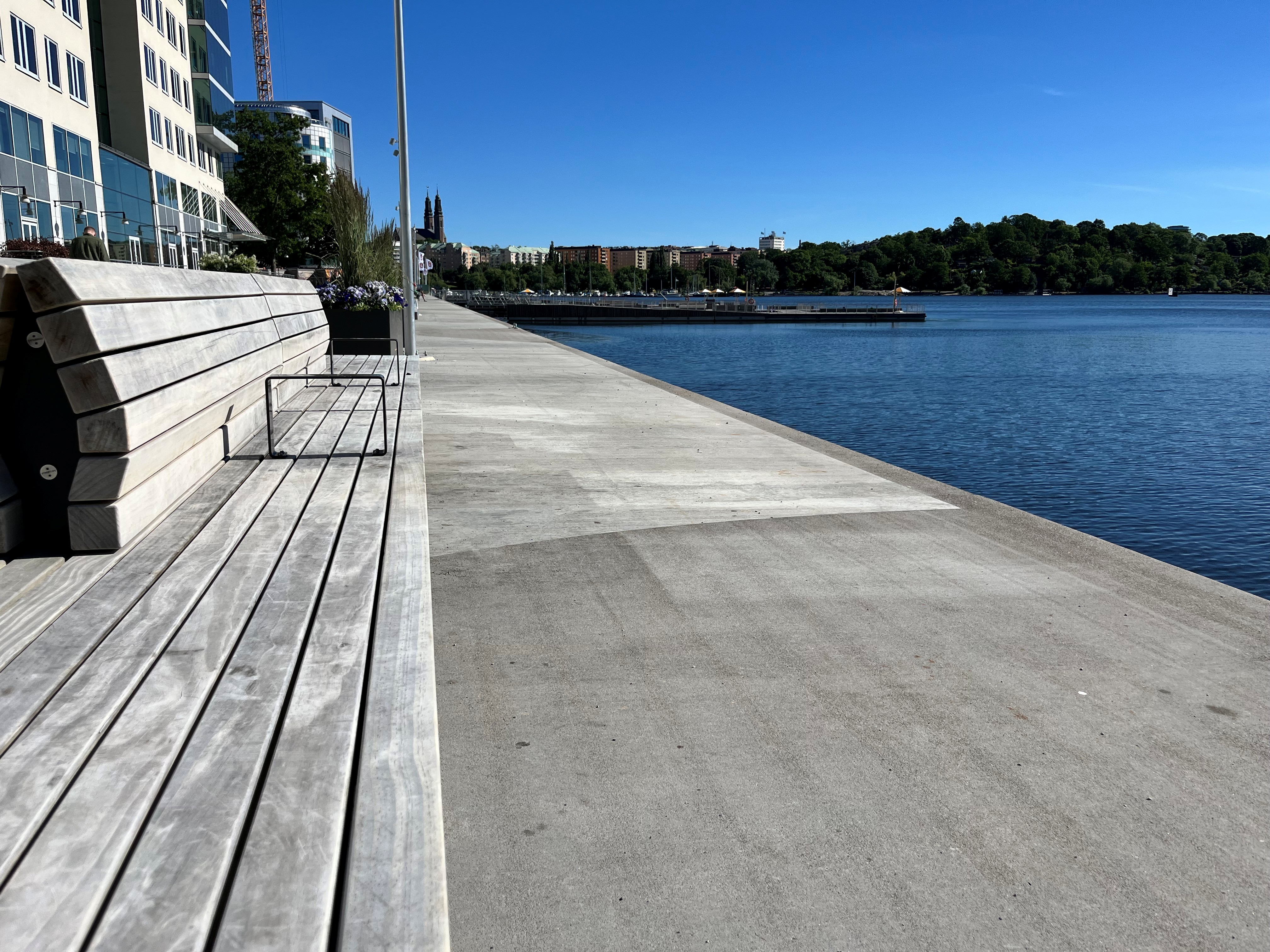 Betongpromenad längs vattnet med träbänkar och byggnader i bakgrunden.