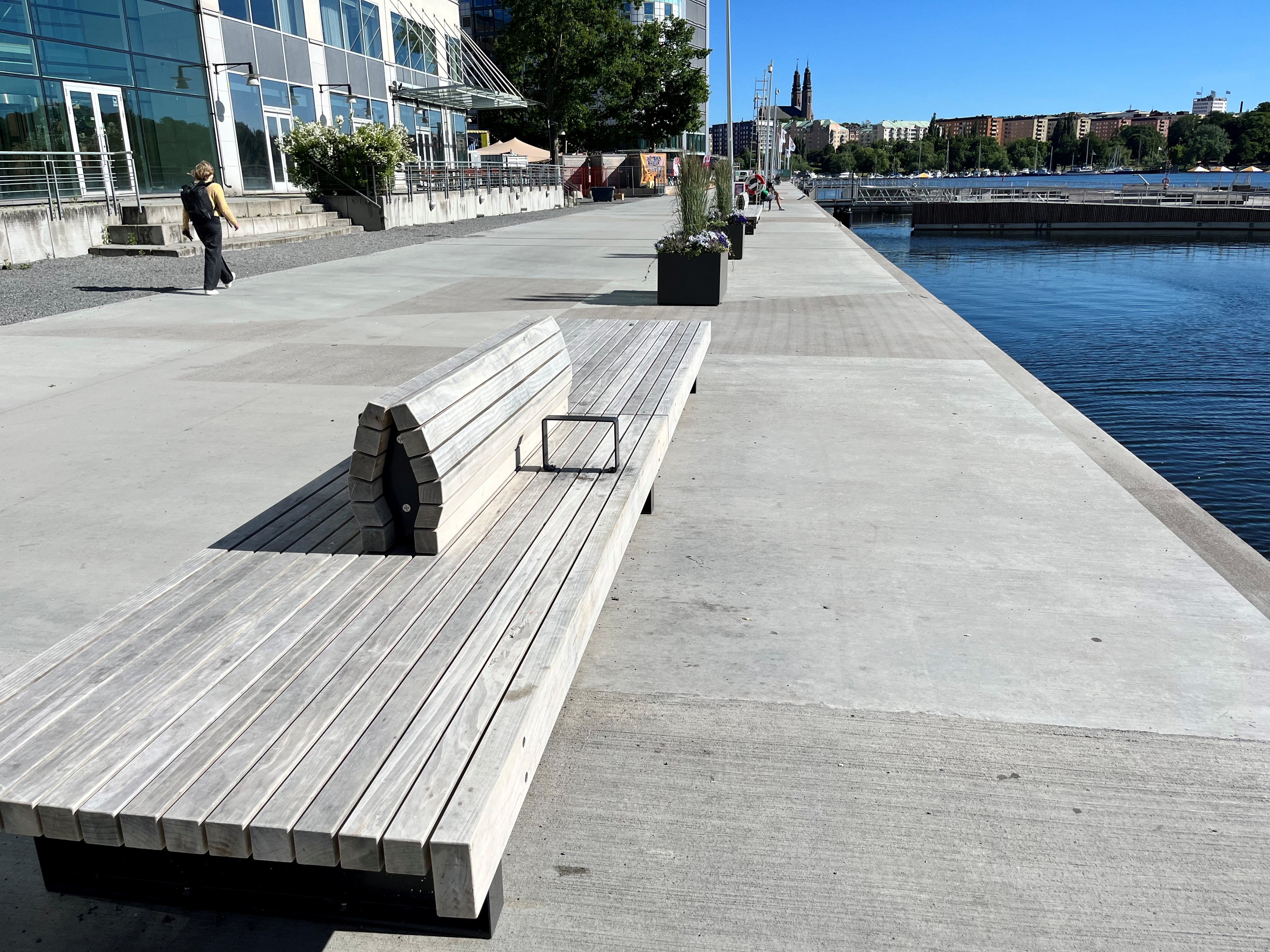 En modern kaj med betongyta, träbänkar och utsikt mot vattnet och byggnader i bakgrunden.