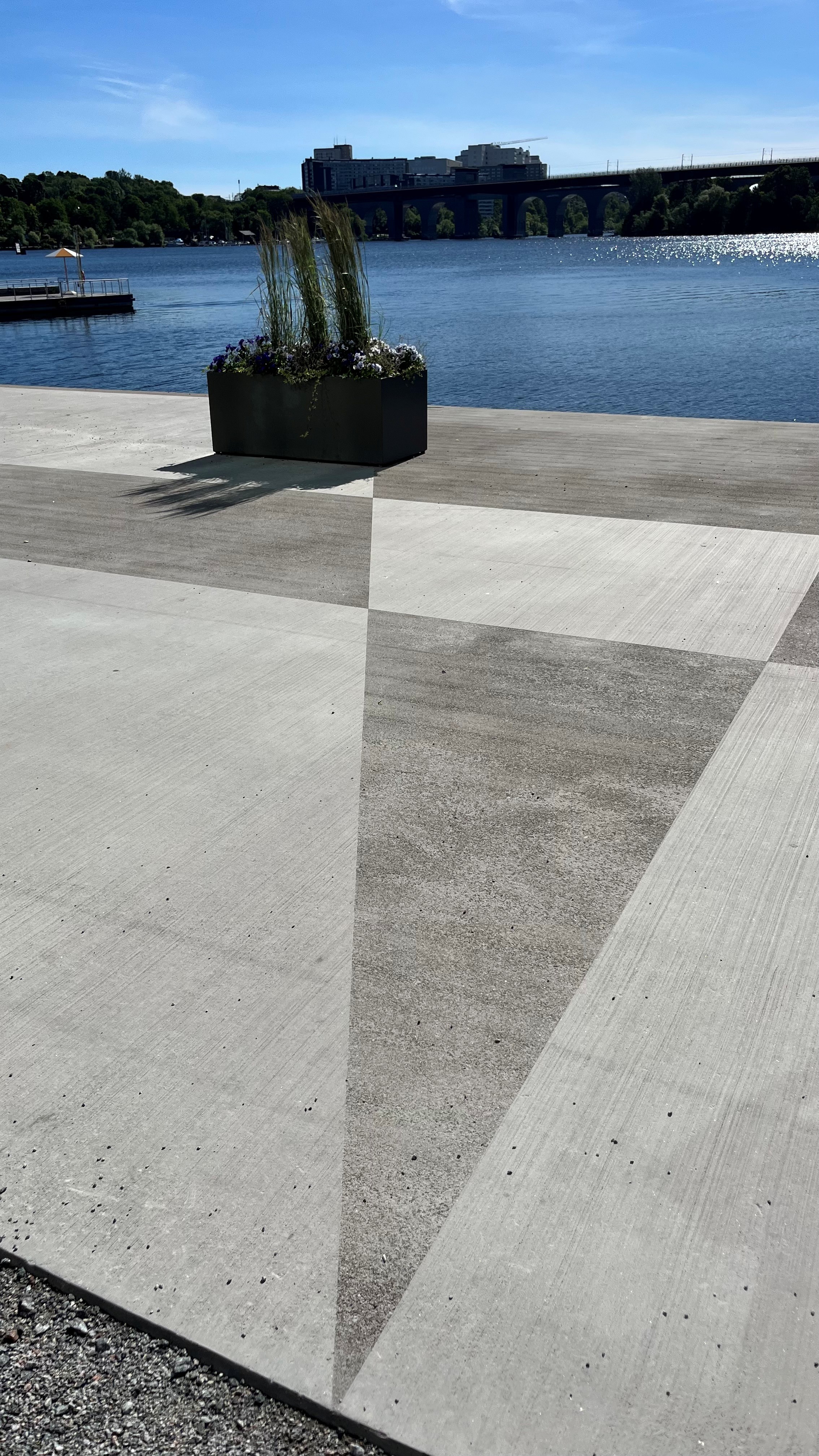 Betongytor vid en vattensida med geometriska mönster och en planteringslåda i bakgrunden.