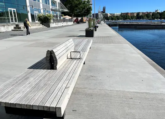 En modern kaj med betongyta, träbänkar och utsikt mot vattnet och byggnader i bakgrunden.